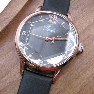 May include: A rose gold wristwatch with a black face and a black band. The watch face has a geometric design with small crystals and the name "Tadi" in the center. The watch is set on a wooden surface.