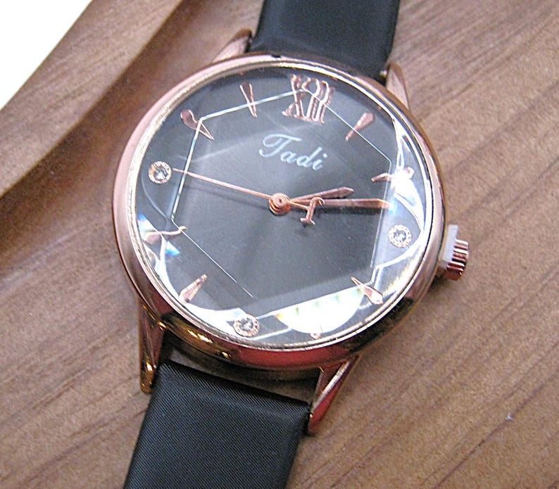 May include: A rose gold wristwatch with a black face and a black band. The watch face has a geometric design with small crystals and the name "Tadi" in the center. The watch is set on a wooden surface.