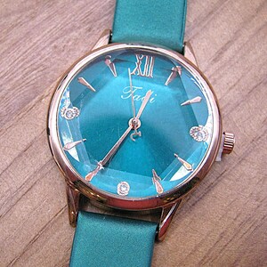 May include: A teal wristwatch with a rose gold case and a matching teal band. The watch face has a teal background with rose gold hands and hour markers. The watch face also has the word "Fadi" in cursive.