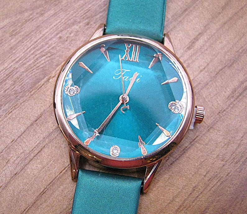 May include: A teal wristwatch with a rose gold case and a matching teal band. The watch face has a teal background with rose gold hands and hour markers. The watch face also has the word "Fadi" in cursive.