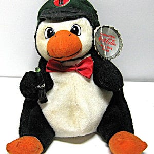 May include: A plush penguin wearing a green Coca-Cola cap and a red bow tie. The penguin holds a miniature Coca-Cola bottle and has a tag shaped like a bottle cap. The penguin has a white chest, black wings, and orange feet and beak.