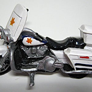 May include: A miniature police motorcycle toy. The motorcycle is primarily white, black, and silver, with orange star decals on the gas tank and windshield. The toy has a windshield, saddlebags, and a black seat.