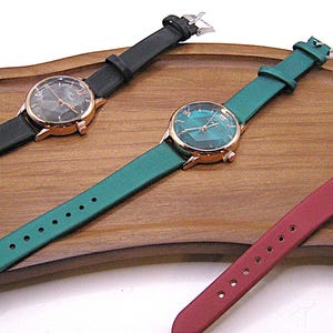 May include: Three wristwatches with leather straps in black, teal, and burgundy. Each watch has a gold-tone case and a patterned face. The watches are displayed on a wooden tray.