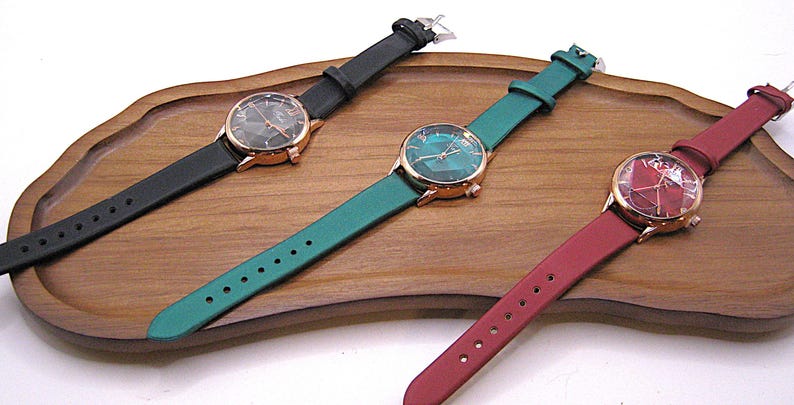 May include: Three wristwatches with leather straps in black, teal, and burgundy. Each watch has a gold-tone case and a patterned face. The watches are displayed on a wooden tray.