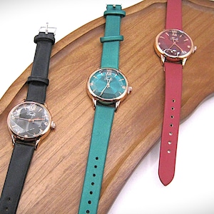 May include: Three elegant wristwatches with gold-tone cases and leather straps in black, teal, and red. The watch faces have a geometric design. The watches are displayed on a wooden tray.