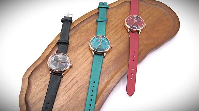 May include: Three elegant wristwatches with gold-tone cases and leather straps in black, teal, and red. The watch faces have a geometric design. The watches are displayed on a wooden tray.
