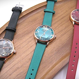 May include: Three elegant wristwatches with rose gold cases and leather straps in black, teal, and burgundy. The watch faces feature geometric designs and are displayed on a wooden tray. The watches are a stylish accessory.