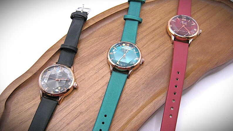 May include: Three elegant wristwatches with rose gold cases and leather straps in black, teal, and burgundy. The watch faces feature geometric designs and are displayed on a wooden tray. The watches are a stylish accessory.