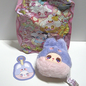 May include: A plush toy with a blue head and pink body, featuring a cartoon face with large eyes. A matching keychain and a pink bag with cartoon characters are also present. The toy has a tag that says "LILATOYS".