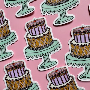 Pu&ograve; includere: Una collezione di adesivi con un design a torta a due piani. Le torte hanno uno schema di colori marrone chiaro e viola, con una ciliegia in cima. Ogni torta poggia su un alzatina verde chiaro con bordo smerlato. Lo sfondo &egrave; rosa.