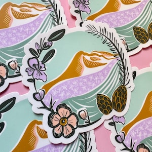 May include: A collection of stickers featuring a nature-themed design. Each sticker has a central image of a mountain range in shades of gold and lavender, surrounded by a floral wreath with pinecones and flowers. The background is a soft teal color.