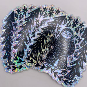May include: A silver glitter sticker featuring a black silhouette of a sloth standing in a forest with trees and flowers. The sticker has a whimsical and cute design.