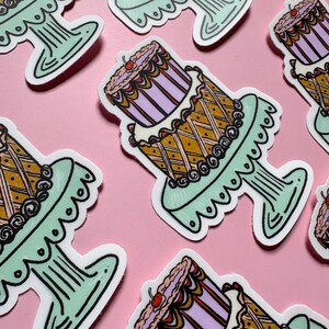 Pu&ograve; includere: Una collezione di adesivi con una torta a due piani su un supporto decorativo. Le torte hanno un design viola e oro, sormontate da una ciliegia. I supporti sono verde chiaro con un bordo smerlato, su uno sfondo rosa.