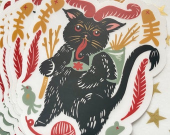 May include: A black cat with red horns and a red tongue is holding a bundle of sticks. The cat is surrounded by red, yellow, and green feathers, a red ball of yarn, and a green mouse. The cat is wearing a green vest with a red flower.