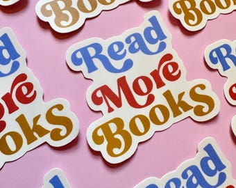 May include: A collection of stickers with the text "Read More Books" in blue, red, and gold lettering. The stickers have a retro font and are arranged on a pink background. The stickers are die-cut with a white border.