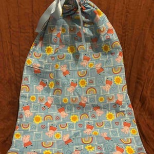 May include: A light blue drawstring bag featuring a repeating pattern of cartoon pigs, rainbows, hearts, and yellow suns. The bag has a light blue ribbon drawstring closure and is made of fabric.