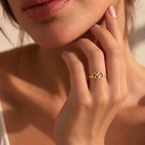Peut inclure: Une bague en or présentant deux contours en forme de cœur entrelacés. L'anneau est fin et circulaire, avec les deux formes de cœur positionnées côte à côte au centre de la bague, créant un design métallique continu, délicat et poli.