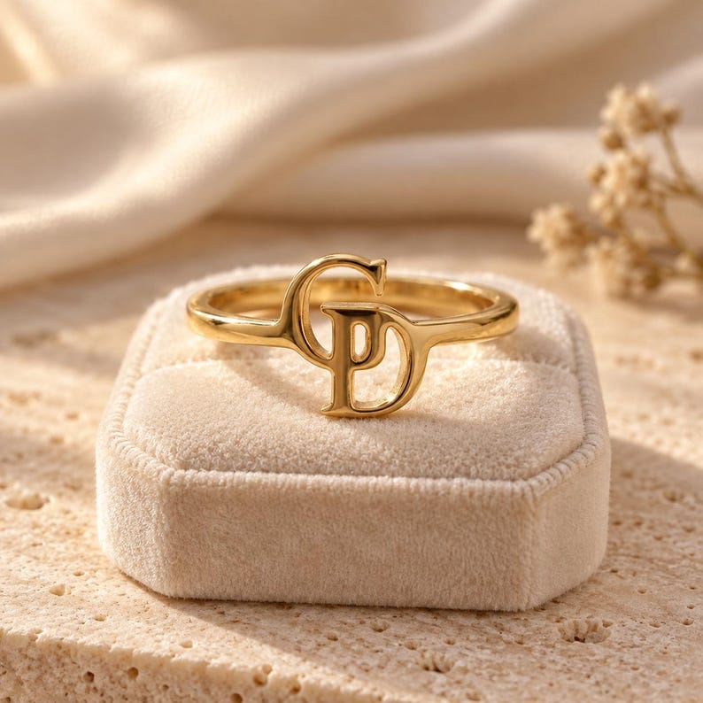 Handcrafted two initial ring in polished gold, 925 sterling silver, and rose gold. Features an elegant interlocking monogram with two custom initials for couples, lovers, or best friends. Dainty personalized letter band worn on woman's finger, displayed on stone tile and velvet ring box. Perfect romantic anniversary gift, promise ring, or birthday gift for her.
