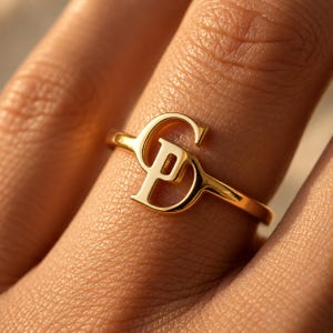 Könnte beinhalten: Ein polierter Goldring mit einem markanten, ineinandergreifenden Monogramm-Design aus den Großbuchstaben S und P. Das Metall hat eine glatte, reflektierende Oberfläche und das Band ist dünn und kreisförmig, wobei die Buchstaben im Mittelpunkt stehen.