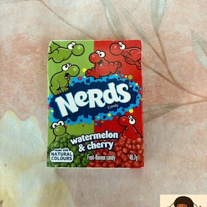 Può includere: Una scatola di caramelle Nerds al gusto di anguria e ciliegia. La scatola è divisa in sezioni verdi e rosse, ognuna con personaggi dei cartoni animati. Il testo "Nerds Candy" è visualizzato, insieme a "realizzato con colori naturali". La scatola contiene 46,7 g di caramelle.
