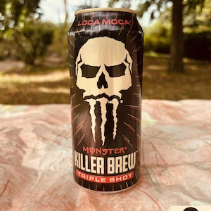 Op de afbeelding: Een zwarte Monster Killer Brew Triple Shot energiedrankblik met een witte schedelafbeelding. Het blik heeft rode accenten en de tekst "Loca Moca" bovenaan. Het blik staat op een patroon.