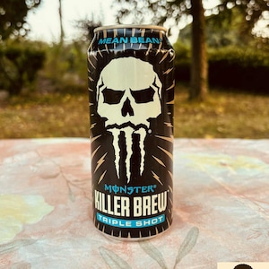 Peut inclure: Une canette noire et dorée de Monster Energy "Mean Bean Killer Brew Triple Shot". La canette présente un graphisme de crâne blanc avec des éclairs. La canette est posée sur une surface à motifs floraux.