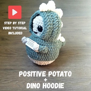 Könnte beinhalten: Ein handgefertigtes gehäkeltes Dinosaurier-Spielzeug in einem tealfarbenen Hoodie. Das Spielzeug hat ein weißes Gesicht und Akzente, mit einem Etikett mit der Aufschrift "Positive Potato". Das Bild enthält auch den Text "Step by Step Video Tutorial Included" und "Positive Potato + Dino Hoodie."
