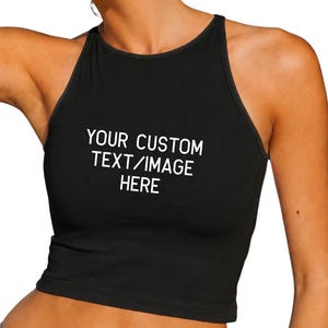 Op de afbeelding: Zwarte halter crop top met de tekst "YOUR CUSTOM TEXT/IMAGE HERE" in het wit. De top heeft een hoge halslijn en een getailleerd silhouet. De stof lijkt een zacht, rekbaar materiaal te zijn.