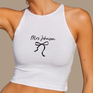 Op de afbeelding: Witte haltertop met de tekst "Mrs. Johnson" in een scriptlettertype en een zwarte strik. De top is mouwloos en heeft een hoge halslijn.