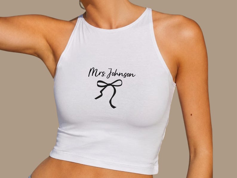 Op de afbeelding: Witte haltertop met de tekst "Mrs. Johnson" in een scriptlettertype en een zwarte strik. De top is mouwloos en heeft een hoge halslijn.