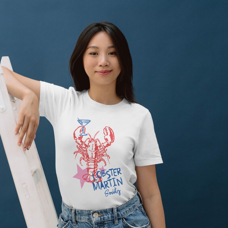 May include: White t-shirt with a red and blue lobster graphic holding a martini glass, and the text "Lobster Martin Society" with a pink star. The shirt is worn with blue jeans.