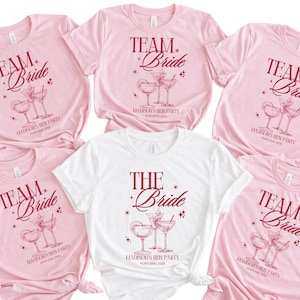 Può includere: Una collezione di magliette rosa e bianche con le scritte "TEAM Bride" e "THE Bride" in bordeaux. Ogni maglietta presenta un disegno di bicchiere da cocktail e la scritta "MADISON'S HEN PARTY PORTOGALLO 2020".