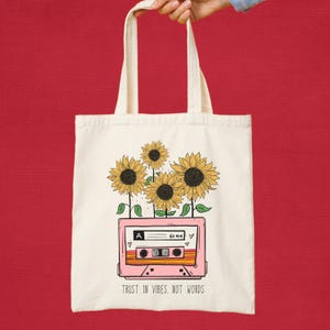 May include: A cream tote bag with a pink cassette tape design and sunflowers. The cassette tape has the text "A 60 MIN" and the phrase "TRUST IN VIBES, NOT WORDS".