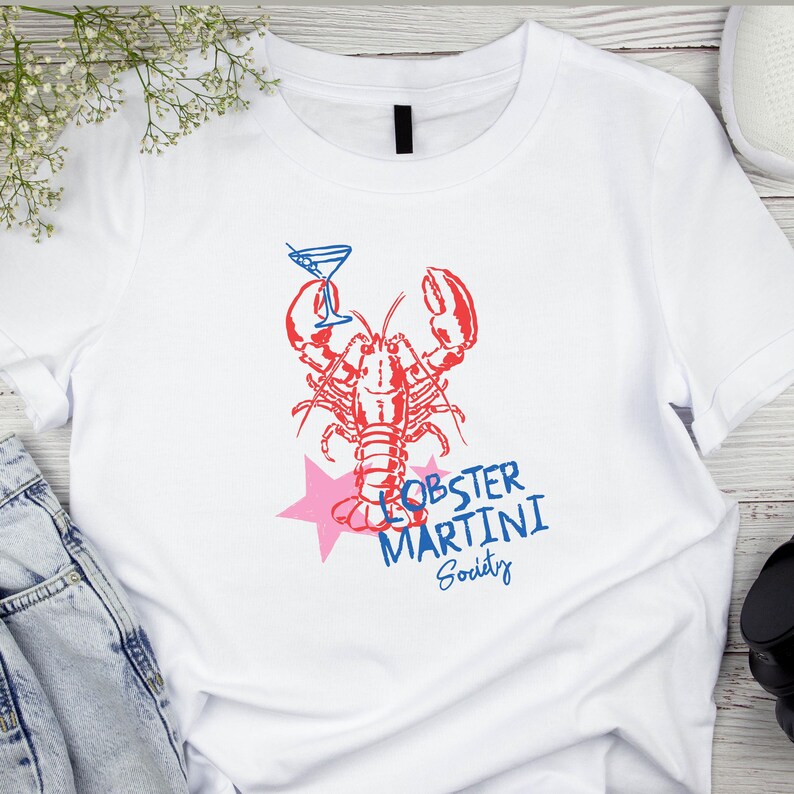 May include: White t-shirt with a red lobster illustration holding a martini. The design includes a pink star and the words "Lobster Martini Society" in blue. A fun, casual design.