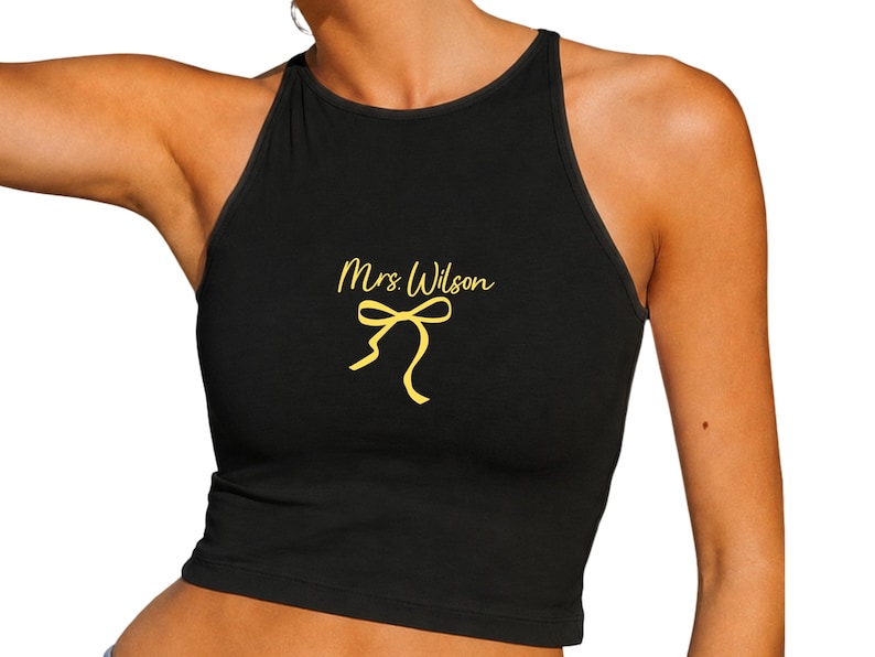 Op de afbeelding: Zwarte haltertop met de tekst "Mrs. Wilson" in een gouden scriptlettertype en een gouden strik. De top is mouwloos en heeft een ronde hals.