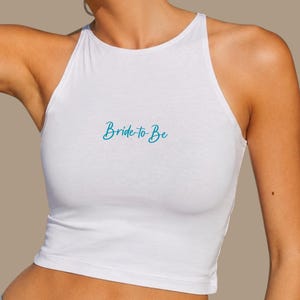 Op de afbeelding: Witte haltertop met de tekst "Bride to Be" in turquoise script. De top is mouwloos en gemaakt van een zacht, lichtgewicht materiaal. De tekst staat in het midden van de voorkant van de top.