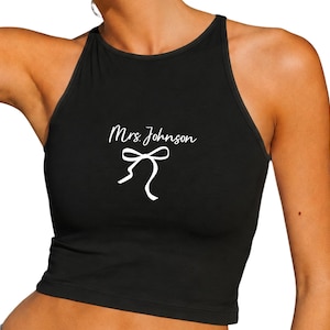 Op de afbeelding: Zwarte haltertop met de tekst "Mrs. Johnson" in cursief en een witte strik. De top is mouwloos en heeft een ronde halslijn.