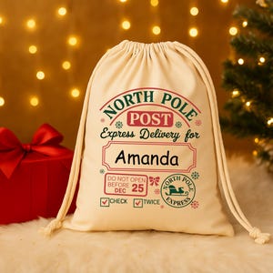 May include: Cream-colored drawstring gift bag with "North Pole Post" and "Express Delivery for Amanda" printed in red and green. Includes a "Do Not Open Before Dec 25" message and a North Pole Express logo.