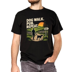 May include: A black short-sleeved t-shirt featuring a graphic print of a person walking a dog on a path. The text reads DOG WALK. PUB. REPEAT. The scene includes a sunset, hills, a stone wall, and a signpost pointing towards a pub.