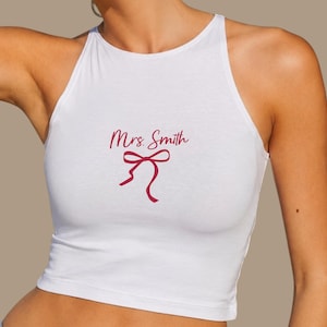 Op de afbeelding: Witte haltertop met de tekst "Mrs. Smith" in een bordeauxrode scriptletter en een bordeauxrode strik. De top is mouwloos en heeft een hoge halslijn.