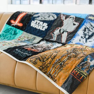 以下が含まれることがあります： リサイクルされたTシャツで作られたスターウォーズをテーマにしたキルト。キルトには、ダース・ベイダー、R2-D2、C-3PO、ミレニアム・ファルコンなど、さまざまなスターウォーズのキャラクターやロゴが描かれています。キルトは茶色の革張りのソファの上に置かれています。