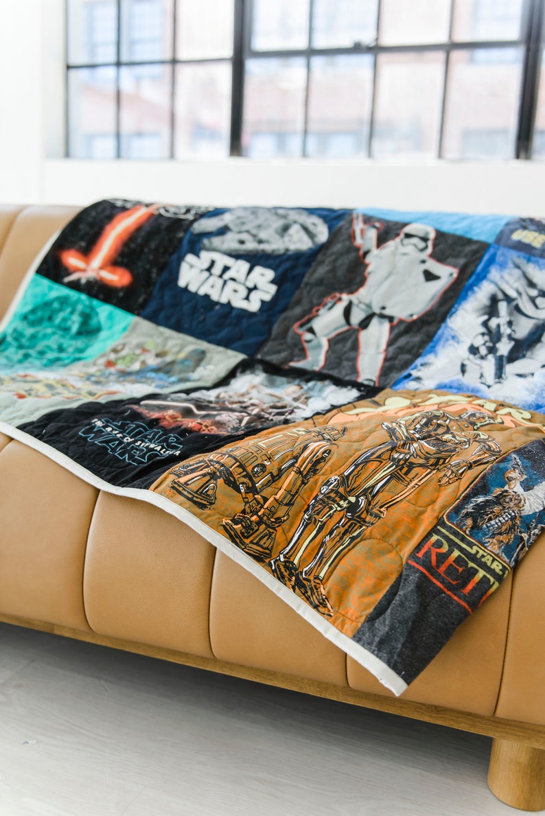 以下が含まれることがあります： リサイクルされたTシャツで作られたスターウォーズをテーマにしたキルト。キルトには、ダース・ベイダー、R2-D2、C-3PO、ミレニアム・ファルコンなど、さまざまなスターウォーズのキャラクターやロゴが描かれています。キルトは茶色の革張りのソファの上に置かれています。
