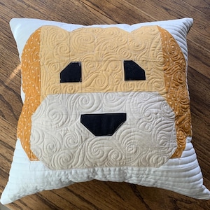 May include: A white pillow with a quilted dog face design. The dog's face is made of beige, brown, and black fabric. The dog's eyes are black and the nose is black.