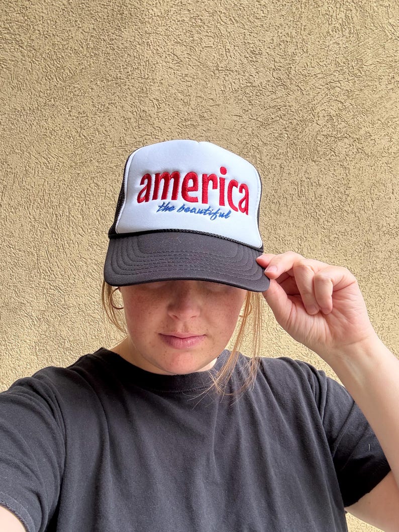 K&ouml;nnte beinhalten: Eine schwarz-wei&szlig;e Trucker-M&uuml;tze mit dem Wort "america" in roter Schrift und "the beautiful" in blauer Schrift. Die M&uuml;tze hat einen schwarzen Schirm und eine wei&szlig;e Frontplatte.