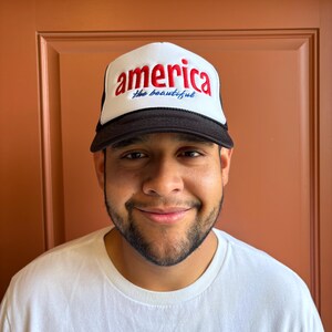 K&ouml;nnte beinhalten: Eine wei&szlig;-schwarze Trucker-M&uuml;tze mit dem Wort "america" in roter Schrift und "the beautiful" in blauer Schrift. Die M&uuml;tze hat einen schwarzen Schirm und eine Netzr&uuml;ckseite. Die M&uuml;tze wird von einer Person getragen, die ein wei&szlig;es T-Shirt tr&auml;gt.