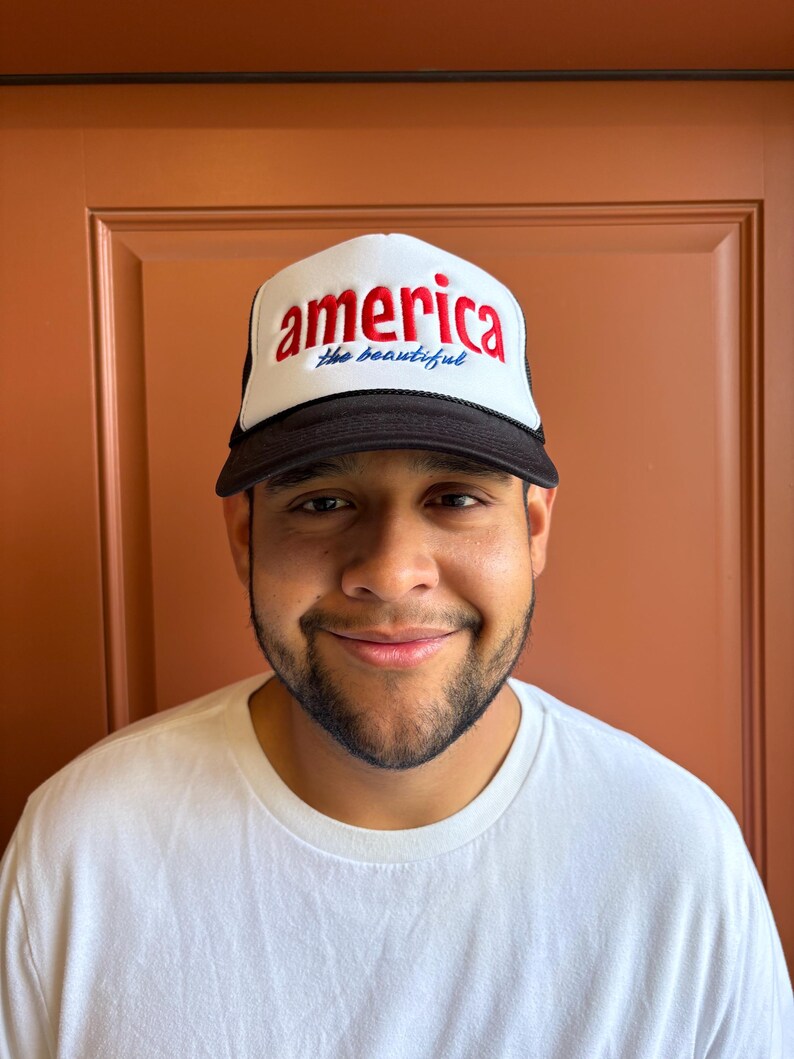 K&ouml;nnte beinhalten: Eine wei&szlig;-schwarze Trucker-M&uuml;tze mit dem Wort "america" in roter Schrift und "the beautiful" in blauer Schrift. Die M&uuml;tze hat einen schwarzen Schirm und eine Netzr&uuml;ckseite. Die M&uuml;tze wird von einer Person getragen, die ein wei&szlig;es T-Shirt tr&auml;gt.