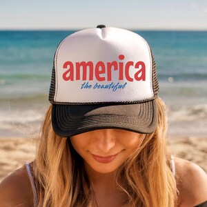 K&ouml;nnte beinhalten: Eine schwarz-wei&szlig;e Trucker-M&uuml;tze mit dem Wort "america" in Rot und "the beautiful" in Blau. Die M&uuml;tze wird von einer Person an einem Strand mit dem Meer im Hintergrund getragen.