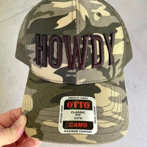 May include: Camouflage baseball cap with the word "HOWDY" embroidered in dark brown. The cap features a mesh back and a curved brim. The front of the cap has a tag that says "OTTO" and "CAMO".