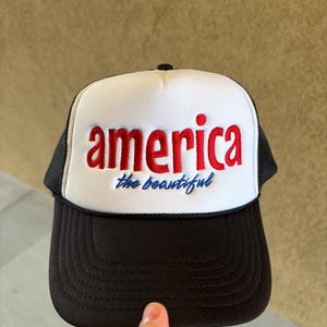 K&ouml;nnte beinhalten: Schwarz-wei&szlig;e Trucker-M&uuml;tze mit dem Wort "america" in roten Blockbuchstaben und dem Satz "the beautiful" in blauer Schrift. Die M&uuml;tze hat einen Mesh-R&uuml;cken und einen gebogenen Schirm.
