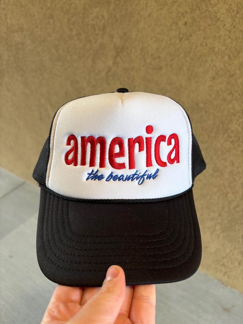 K&ouml;nnte beinhalten: Schwarz-wei&szlig;e Trucker-M&uuml;tze mit dem Wort "america" in roten Blockbuchstaben und dem Satz "the beautiful" in blauer Schrift. Die M&uuml;tze hat einen Mesh-R&uuml;cken und einen gebogenen Schirm.